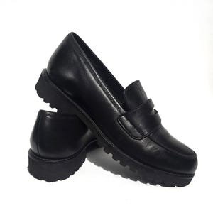 May include: A pair of black leather loafers with a chunky, ridged sole. The shoes have a classic penny loafer design with a strap across the vamp. The leather appears smooth and the shoes are set against a white background.