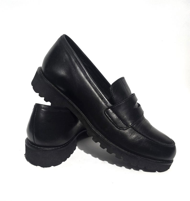 May include: A pair of black leather loafers with a chunky, ridged sole. The shoes have a classic penny loafer design with a strap across the vamp. The leather appears smooth and the shoes are set against a white background.