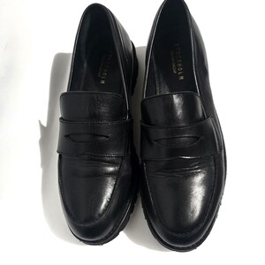 May include: A pair of black leather loafers with a rounded toe and a strap across the top. The shoes have a thick sole and the word "STOCKHOLM" is printed on the side.