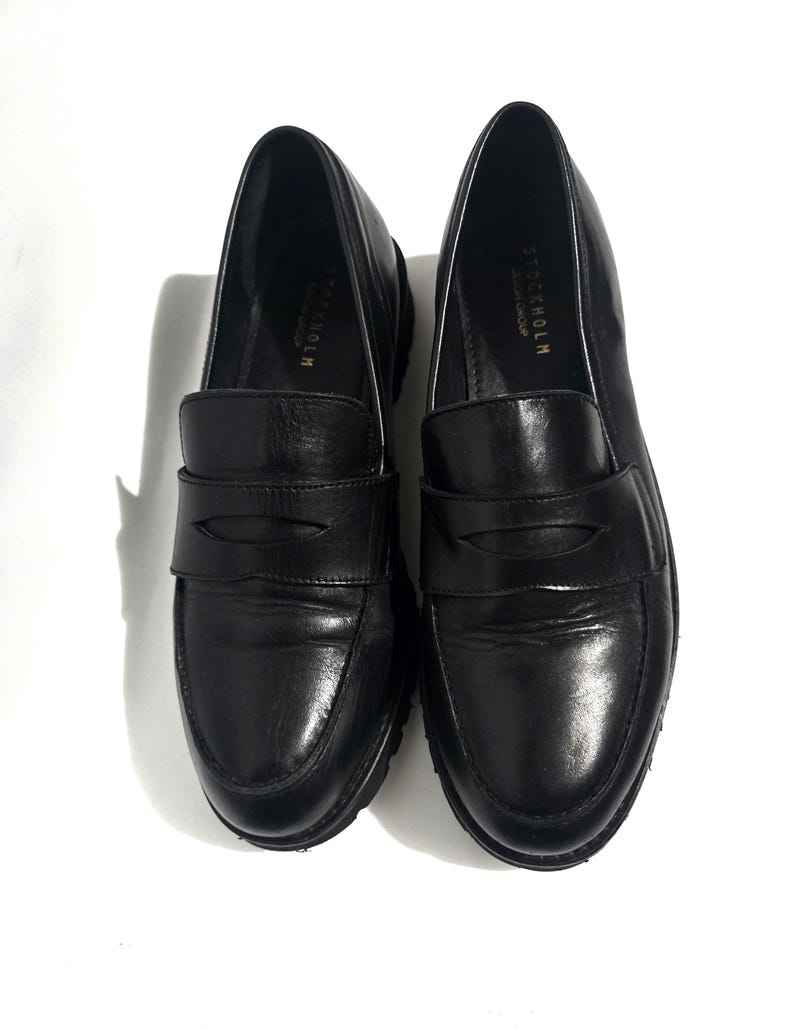 May include: A pair of black leather loafers with a rounded toe and a strap across the top. The shoes have a thick sole and the word "STOCKHOLM" is printed on the side.