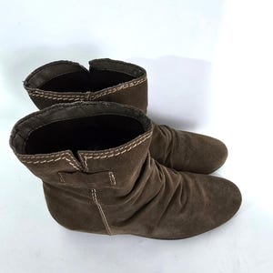May include: A pair of brown suede ankle boots. The boots have a rounded toe and a low heel. The top of the boots are folded over and have white stitching. The boots are made of a soft, suede material.