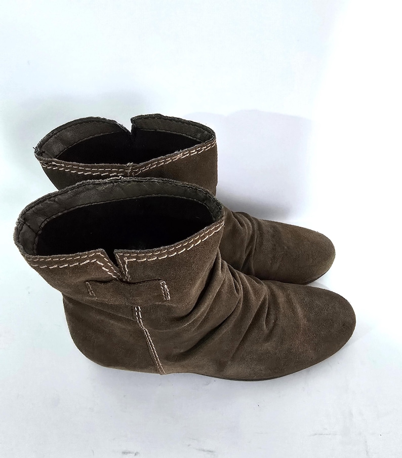 May include: A pair of brown suede ankle boots. The boots have a rounded toe and a low heel. The top of the boots are folded over and have white stitching. The boots are made of a soft, suede material.