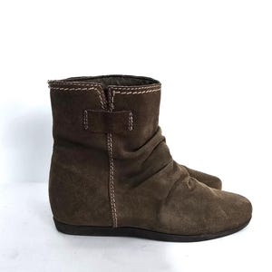 May include: Brown suede ankle boots with a low heel. The boots feature a ruched design on the upper and a small buckle detail. White stitching accents the seams and the top of the boot.