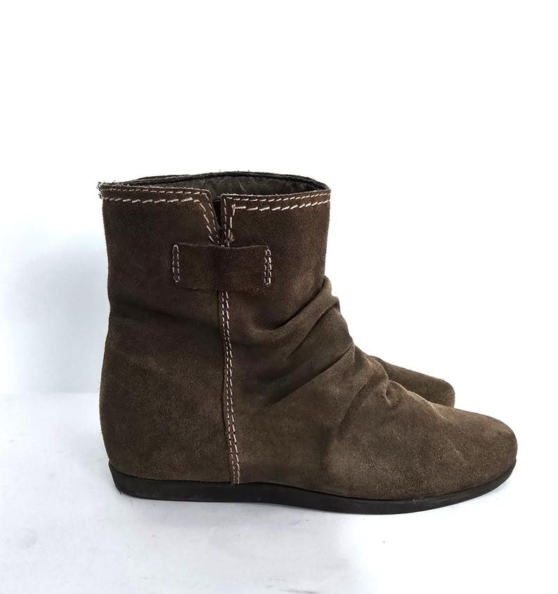 May include: Brown suede ankle boots with a low heel. The boots feature a ruched design on the upper and a small buckle detail. White stitching accents the seams and the top of the boot.