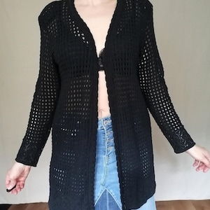 May include: A black open-front cardigan featuring a long-sleeve design with a repeating grid-like crochet pattern. The garment has a scalloped edge along the front opening and neckline, extending to a mid-thigh length with a lightweight, airy texture.