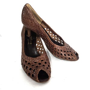 May include: A pair of brown peep-toe heels with a snakeskin pattern. The shoes have a cut-out design on the upper and a small heel. The shoes are set against a white background.