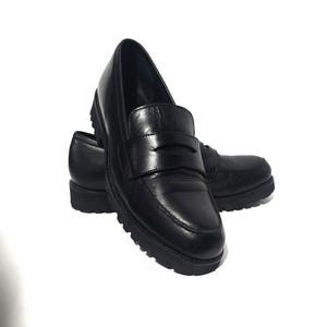 May include: A pair of black leather penny loafers with a chunky sole. The shoes have a classic design with a strap across the vamp. The soles are thick and have a textured pattern for added grip. The shoes are set against a white background.