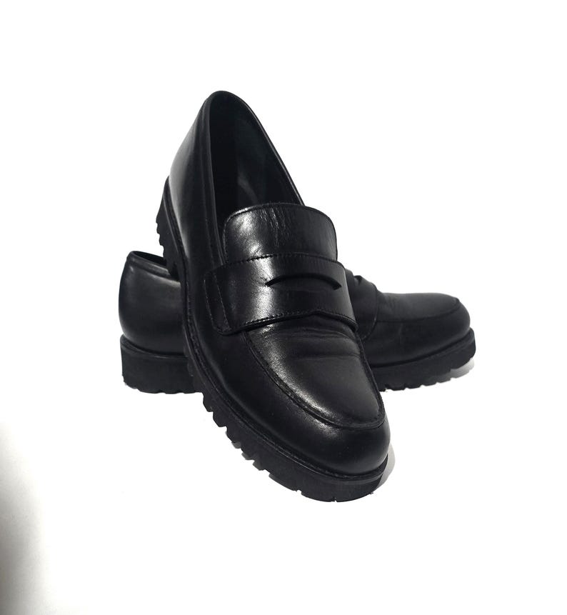 May include: A pair of black leather penny loafers with a chunky sole. The shoes have a classic design with a strap across the vamp. The soles are thick and have a textured pattern for added grip. The shoes are set against a white background.