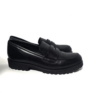 May include: A pair of black leather loafers with a rounded toe and a thick, textured sole. The shoes have a slip-on design with a strap across the top. The loafers are displayed on a white background.