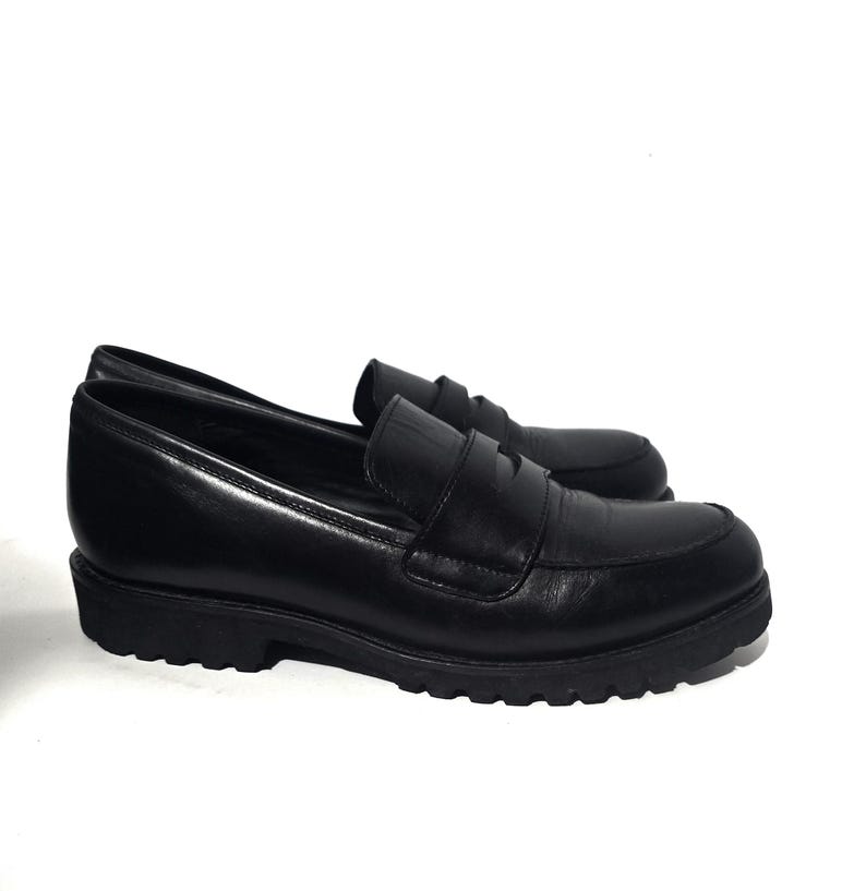 May include: A pair of black leather loafers with a rounded toe and a thick, textured sole. The shoes have a slip-on design with a strap across the top. The loafers are displayed on a white background.