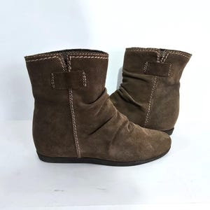May include: A pair of brown suede ankle boots. The boots have a low heel, a ruched design, and a strap detail with a buckle. White stitching accents the edges and seams of the boots.