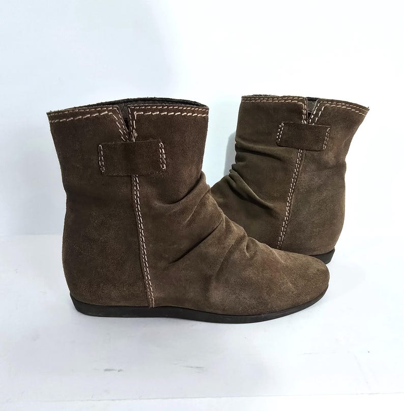 May include: A pair of brown suede ankle boots. The boots have a low heel, a ruched design, and a strap detail with a buckle. White stitching accents the edges and seams of the boots.