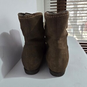 May include: A pair of brown suede ankle boots. The boots have a rounded toe and a short heel. The top of the boots are stitched with a contrasting thread. The boots are displayed on a white surface.