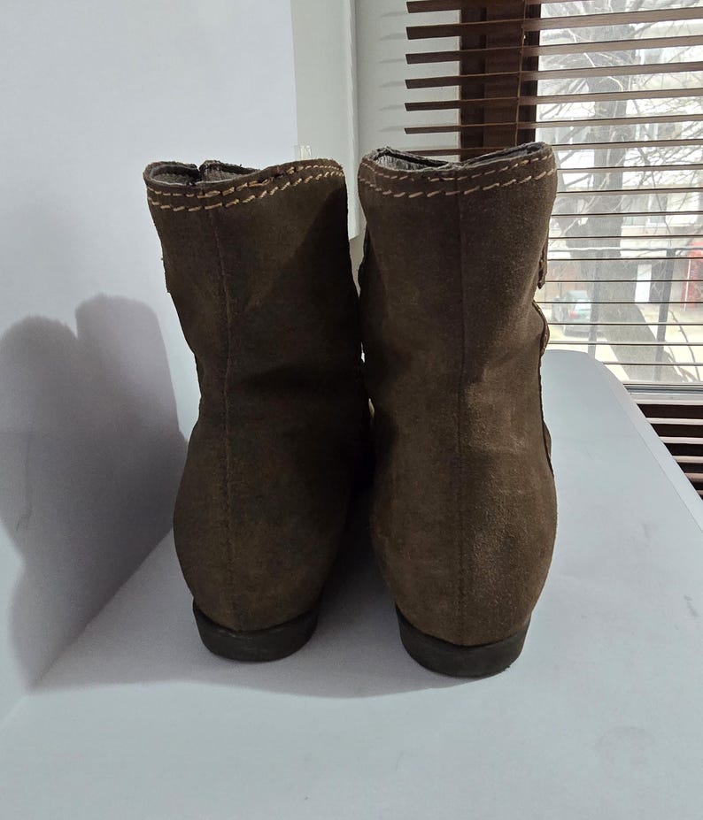 May include: A pair of brown suede ankle boots. The boots have a rounded toe and a short heel. The top of the boots are stitched with a contrasting thread. The boots are displayed on a white surface.