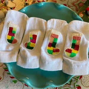 May include: Four white socks with a painted Christmas stocking design on each. The stockings are painted with red, green, yellow, and pink squares.