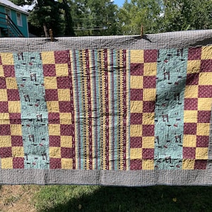 Może przedstawiać: Kołdra w kratę w kolorze żółtym i bordowym, w pasy w kolorze niebieskim i żółtym oraz z wzorzystą tkaniną na niebieskim tle. Kołdra jest powieszona na sznurze do suszenia.