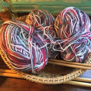 Puede incluir: Tres ovillos de lana en una cesta de mimbre. La lana es una mezcla de rosa, blanco, gris y rojo.