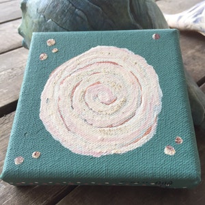 May include: A small, teal canvas painting with a white and pink spiral design in the centre. The spiral is surrounded by small brown dots.