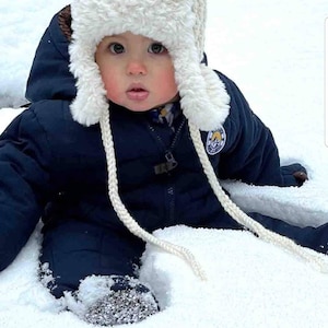 May include: A baby wearing a navy blue snowsuit and a white, fluffy trapper hat with braided ties, sitting in the snow. The hat has ear flaps and a brown interior. The baby's face is visible, with rosy cheeks and a curious expression.