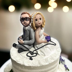May include: A white wedding cake with a purple and white floral design. On top of the cake is a fondant couple holding video game controllers.