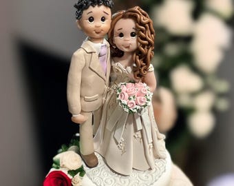 May include: A white wedding cake with a fondant bride and groom topper. The bride is wearing a white dress with pink flowers and the groom is wearing a tan suit. The cake is decorated with white frosting and a lace design.