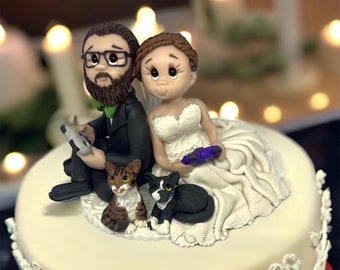 Könnte beinhalten: Eine weiße Hochzeitstorte mit einer Fondant-Braut- und Bräutigamfigur, einer braun-weißen Katze und einer schwarz-weißen Katze. Die Braut trägt ein weißes Kleid und hält einen violetten Videospiel-Controller. Der Bräutigam trägt einen schwarzen Anzug und hält einen Videospiel-Controller. Die Torte ist mit weißer Spitze verziert.