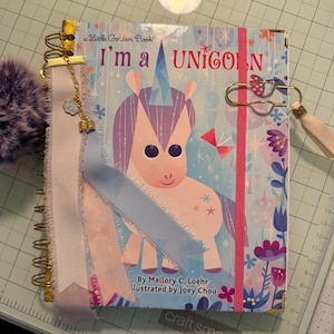 Può includere: Un taccuino a spirale con la copertina del libro Little Golden Book I'm a Unicorn di Mallory C. Loehr. La copertina mostra un unicorno, motivi floreali e un elastico rosa. Include una spirale dorata, una nappa di nastro con un ciondolo a forma di libro e una grande graffetta dorata con una nappa bianca.
