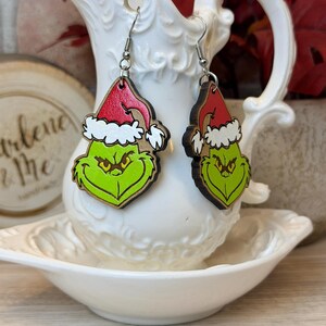May include: A pair of wooden dangle earrings featuring the Grinch's face, complete with a red Santa hat and a green face. The earrings are hanging in front of a white decorative pitcher.