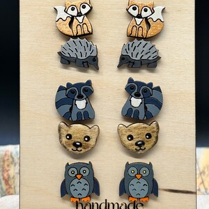 May include: A set of six pairs of wooden earrings featuring various woodland animals. The earrings are painted in shades of brown, gray, and blue. The animals include foxes, hedgehogs, raccoons, bears, and owls. The earrings are displayed on a wooden board with the word "handmade" printed on it.