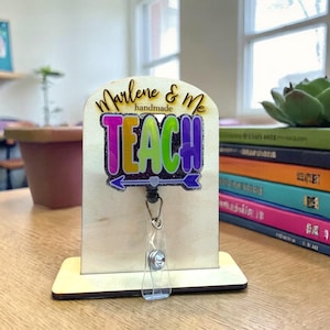 Può includere: Un porta badge in legno con la scritta "Marlene & Me handmade TEACH" in lettere colorate. Il porta badge ha un avvolgitore retrattile in plastica trasparente ed è esposto su un supporto in legno. Una pila di libri e una pianta succulenta sono sullo sfondo.