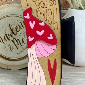 Könnte beinhalten: Rechteckige Holzdekoration mit einem Pilzdesign mit rotem Hut und rosa Herzen. Der Text "I love you so mush" befindet sich über dem Pilz. Unten befindet sich ein rotes Herz.