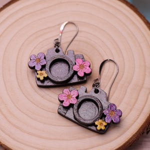 Puede incluir: Un par de pendientes de plata con un diseño de cámara y detalles florales. La cámara está hecha de madera y pintada de negro con un acabado de purpurina plateada. Las flores son rosas, moradas y amarillas.
