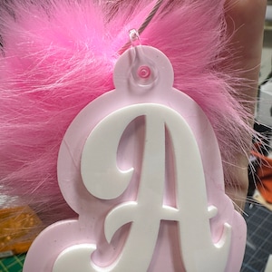 Könnte beinhalten: Ein rosa-weißes Schlüsselanhänger mit Monogramm und einem flauschigen rosa Federakzent. Der Schlüsselanhänger hat eine hellrosa Basis mit dem weißen Buchstaben "A" in Schreibschrift. Eine silberfarbene Drahtschlaufe ist befestigt.