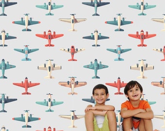May include: A white wall with a repeating pattern of colourful aeroplanes. Two children are sitting in front of the wall. The children are wearing casual clothing and smiling.