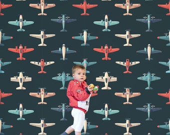 May include: A young child wearing a red jacket and white pants stands in front of a wall covered in a repeating pattern of colorful airplanes. The child is holding a small toy airplane in each hand.