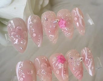 May include: A set of ten almond-shaped artificial nails. Each nail is painted a light pink with a glossy finish. Some nails feature small, clear embellishments, while others have pink flower accents and clear gems. The nails are displayed on a clear surface.