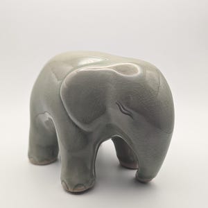 May include: A ceramic elephant figurine in a soft, muted green colour. The elephant has a smooth, glossy finish with a crackle glaze effect. The figurine is standing with its trunk down and its ears slightly flared.
