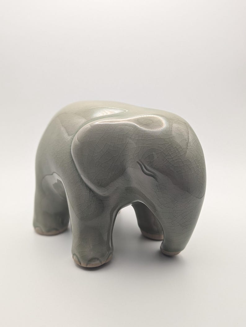 May include: A ceramic elephant figurine in a soft, muted green colour. The elephant has a smooth, glossy finish with a crackle glaze effect. The figurine is standing with its trunk down and its ears slightly flared.