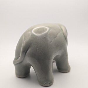 May include: A ceramic elephant ornament in a muted green-grey hue. The elephant is shown in a downward posture, with its trunk touching the ground. The figurine has a textured, crackled surface and is set against a white backdrop.
