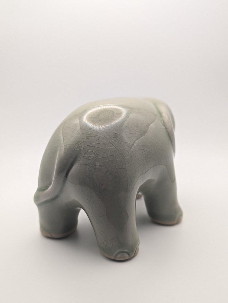 May include: A ceramic elephant ornament in a muted green-grey hue. The elephant is shown in a downward posture, with its trunk touching the ground. The figurine has a textured, crackled surface and is set against a white backdrop.