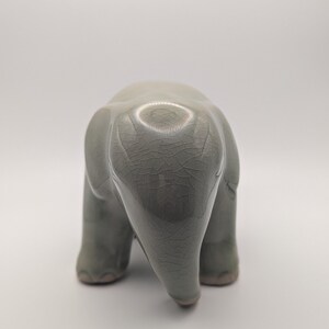 May include: A ceramic elephant figurine in a muted green-grey glaze. The elephant is viewed from the rear, showing its rounded form and textured surface. The figurine has a crackle finish, adding to its unique aesthetic.