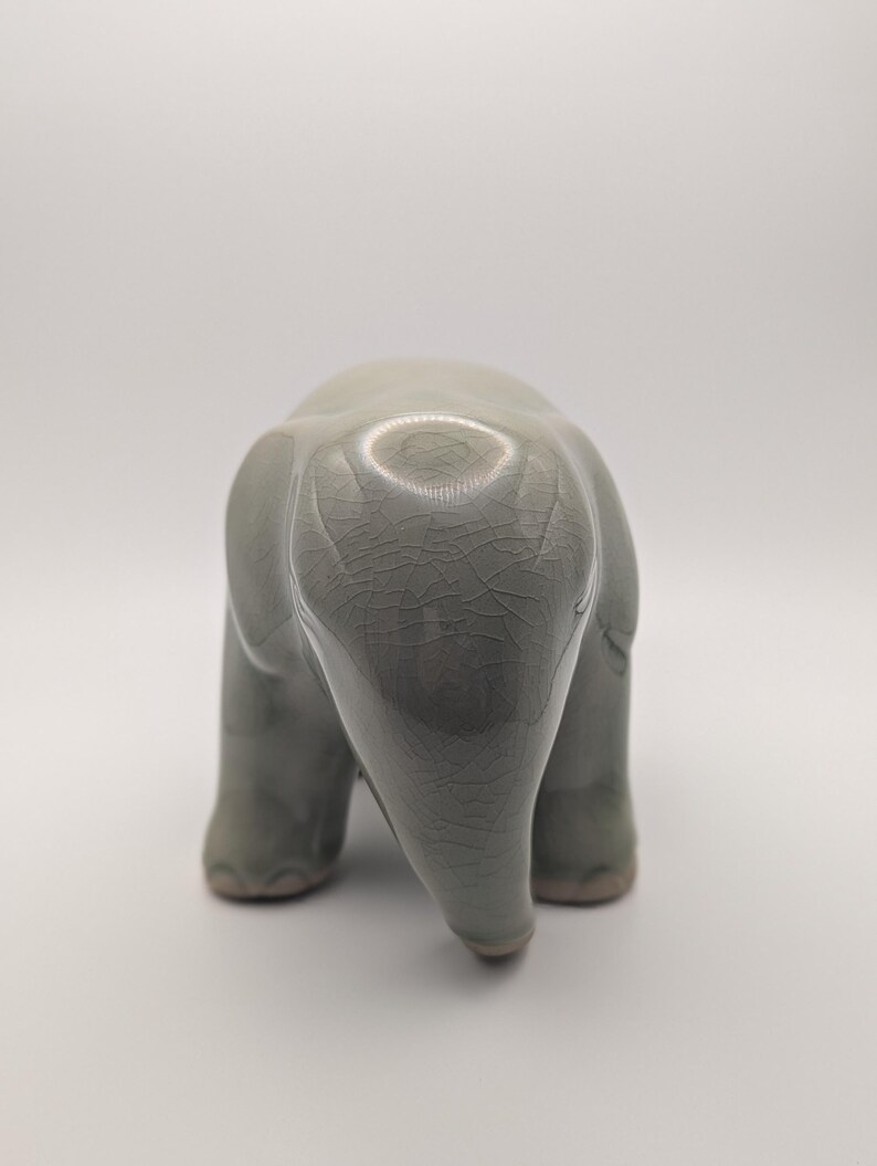 May include: A ceramic elephant figurine in a muted green-grey glaze. The elephant is viewed from the rear, showing its rounded form and textured surface. The figurine has a crackle finish, adding to its unique aesthetic.