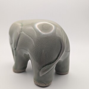 May include: A ceramic elephant figurine in a muted green-grey colour. The elephant is depicted from the rear, showcasing its textured skin and rounded form. The figurine has a glossy finish and is set against a white background.