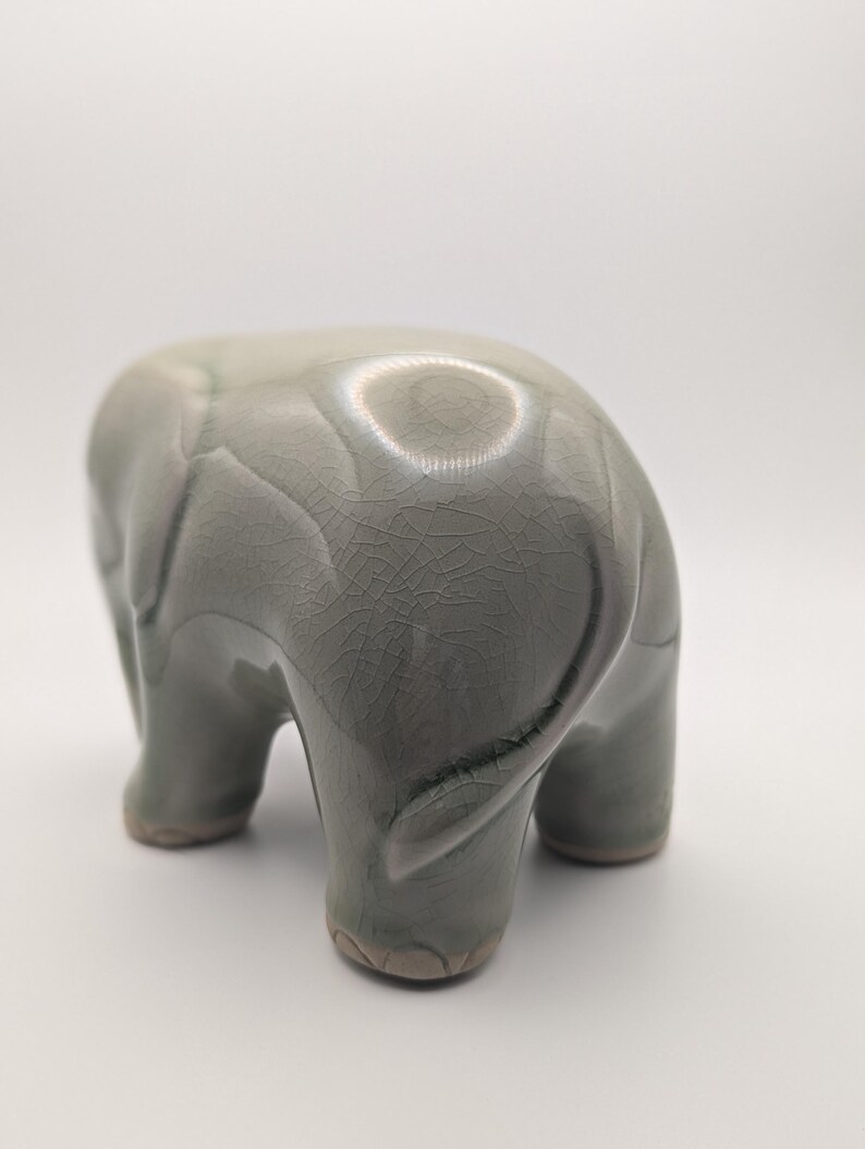 May include: A ceramic elephant figurine in a muted green-grey colour. The elephant is depicted from the rear, showcasing its textured skin and rounded form. The figurine has a glossy finish and is set against a white background.