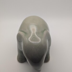 May include: A celadon ceramic elephant figurine. The elephant is depicted in a stylized form, with a smooth, rounded body and visible cracks in the glaze. The figurine has a subtle gradient of colour, with darker tones at the base.