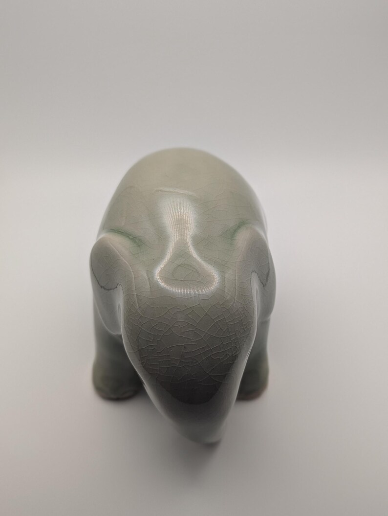 May include: A celadon ceramic elephant figurine. The elephant is depicted in a stylized form, with a smooth, rounded body and visible cracks in the glaze. The figurine has a subtle gradient of colour, with darker tones at the base.