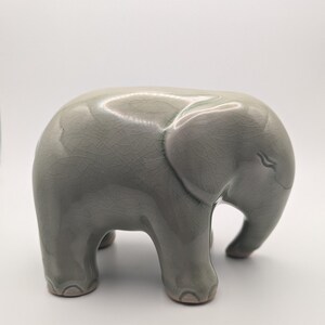 May include: A light grey ceramic elephant figurine. The elephant has a smooth, glossy finish with a crackle pattern. The figurine is standing on four short legs, with its trunk curved downwards. A decorative item, suitable for display.