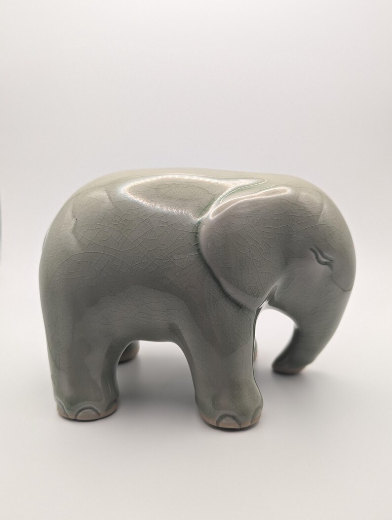 May include: A light grey ceramic elephant figurine. The elephant has a smooth, glossy finish with a crackle pattern. The figurine is standing on four short legs, with its trunk curved downwards. A decorative item, suitable for display.
