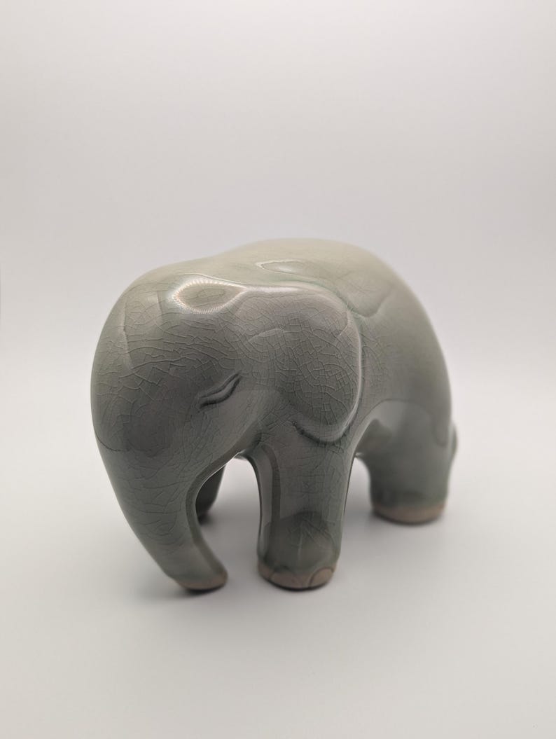May include: A grey ceramic elephant ornament with a crackle glaze. The elephant has a rounded body, small ears, and a downward-curving trunk. The figurine is approximately 15 cm long and 10 cm tall.
