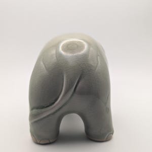 May include: A light grey-green ceramic elephant figurine, seen from behind. The elephant has a textured, crackled surface and a curled tail. The figurine has a rounded body and stands on four legs. The measurements are in centimetres.